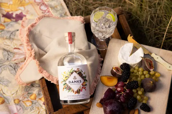 Banks Botanicals Bottle with cheese board and fruit laid on picnic rung