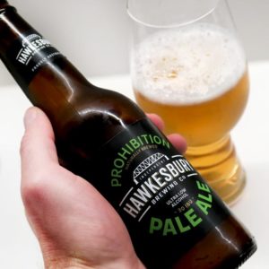 Hawkesbury Pale Ale bottle in hand with glass behind it
