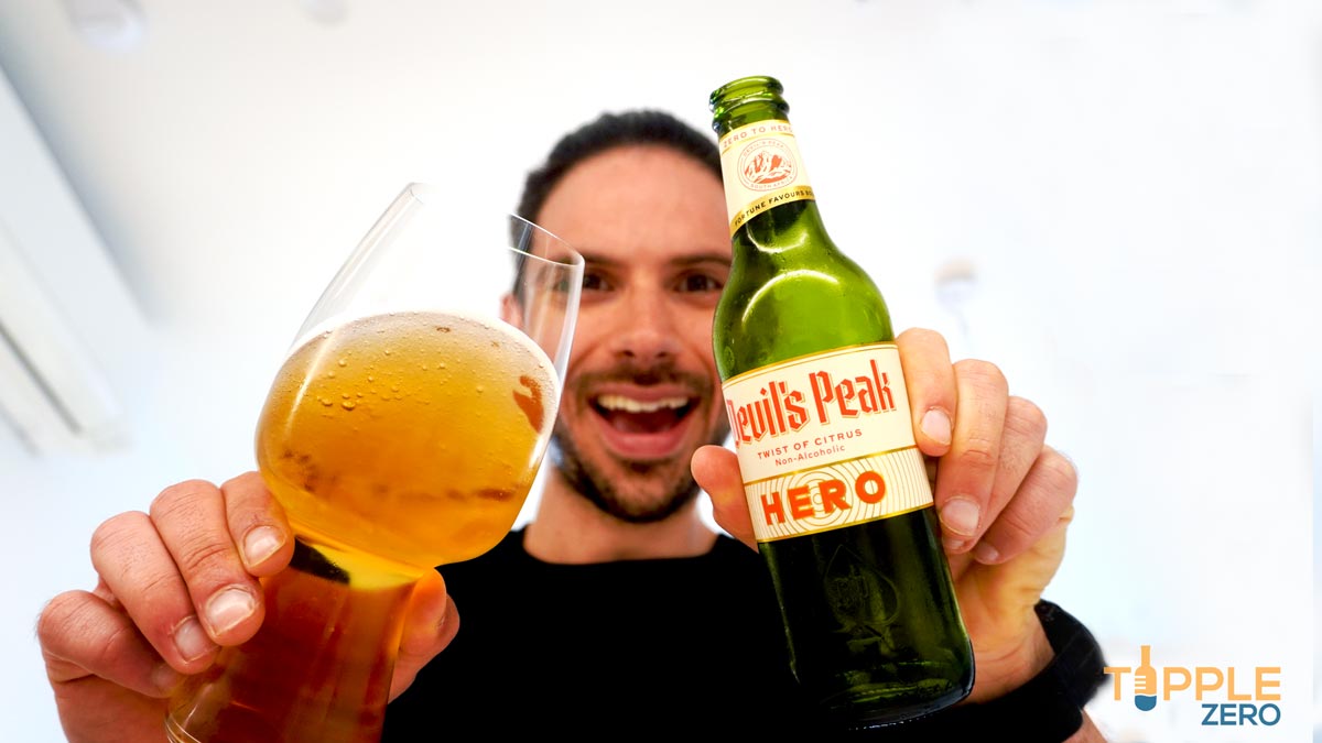 Devils Peak Hero Twist of Citrus held in hand next to glass of non-alcoholic beer